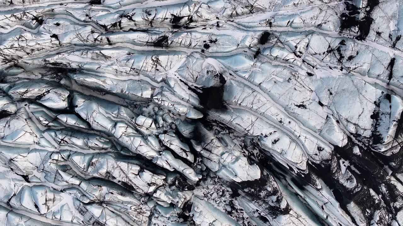 Top-down aerial view of the Sólheimajökull glacier, an outlet of the Mýrdalsjökull ice cap in Iceland. Rugged ice formations, blue tones, and streaks of volcanic ash create a abstract landscape.