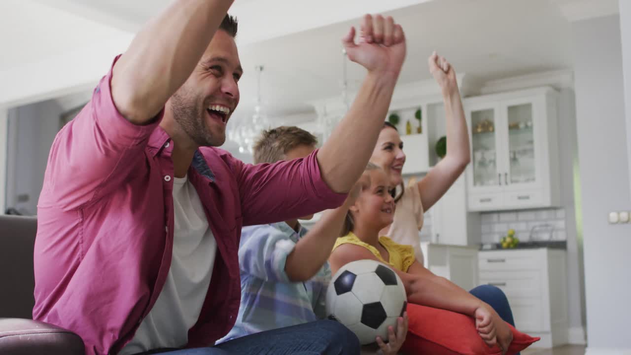 padres caucásicos emocionados, hijo e hija en el sofá viendo televisión y animando, hijo sosteniendo un fútbol