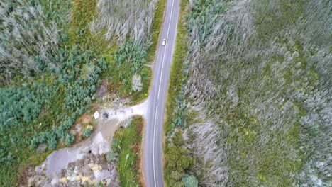 Aerial-shot-looking-directly-down-at-a-white-car,-following-it-as-it-drives-along-through-forest