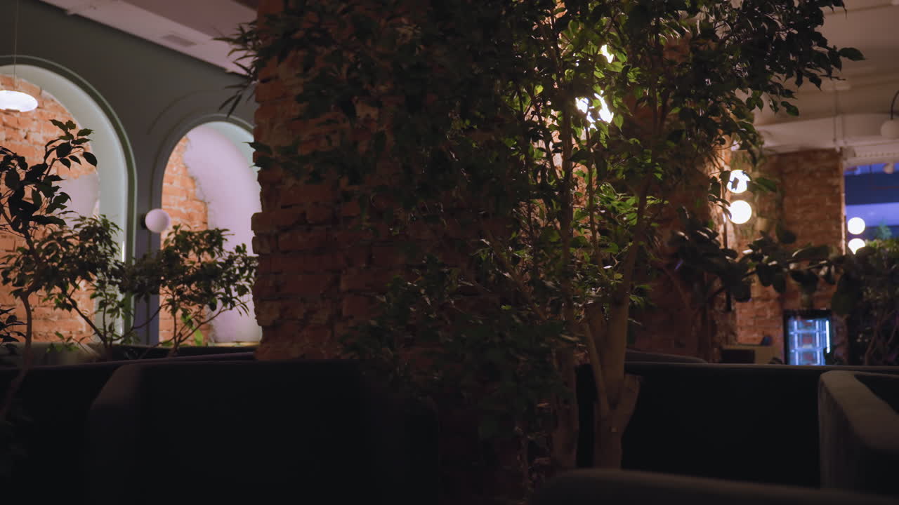 Room interior features exposed brick walls, indoor plants, and comfortable cushioned seats. Soft lighting and lush greenery create relaxing natural atmosphere enhanced by architectural elements