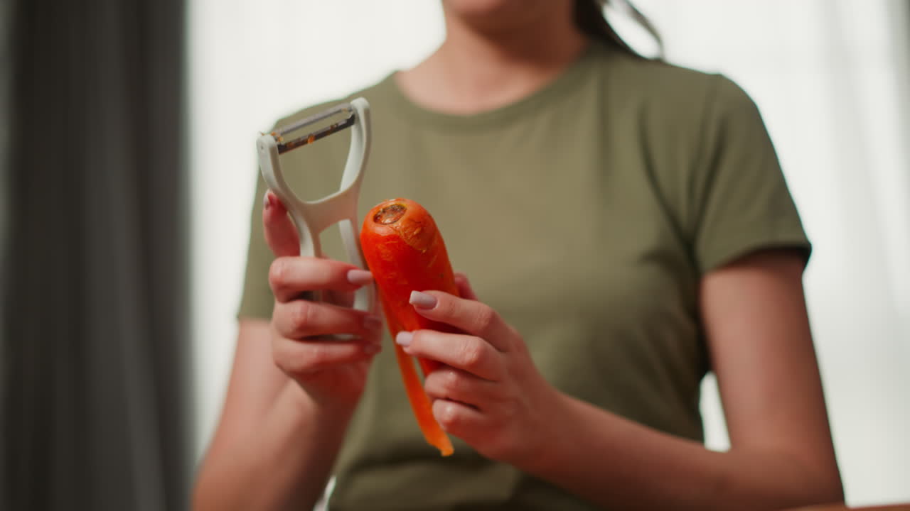 Close up of woman holding fresh carrot and using handheld peeler to remove outer skin. Background softly blurred with focus on carrot, hands, and peeler movement during vegetable preparation