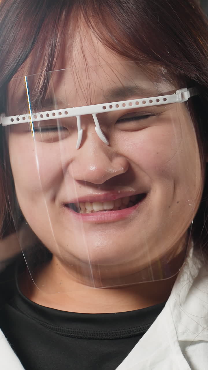 보호 얼굴 방패를 착용 한 학생은 기계 작업장에서 따뜻한 미소로 눈을 니다. 배경에는 산업 도구, 밝은 조명 및 기계가 있습니다.