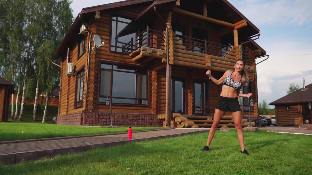 una mujer delgada y hermosa con ropa deportiva con una prensa abierta se está preparando para comenzar a entrenar en el césped cerca de su casa realizando pendientes flexionando los músculos de la espalda
