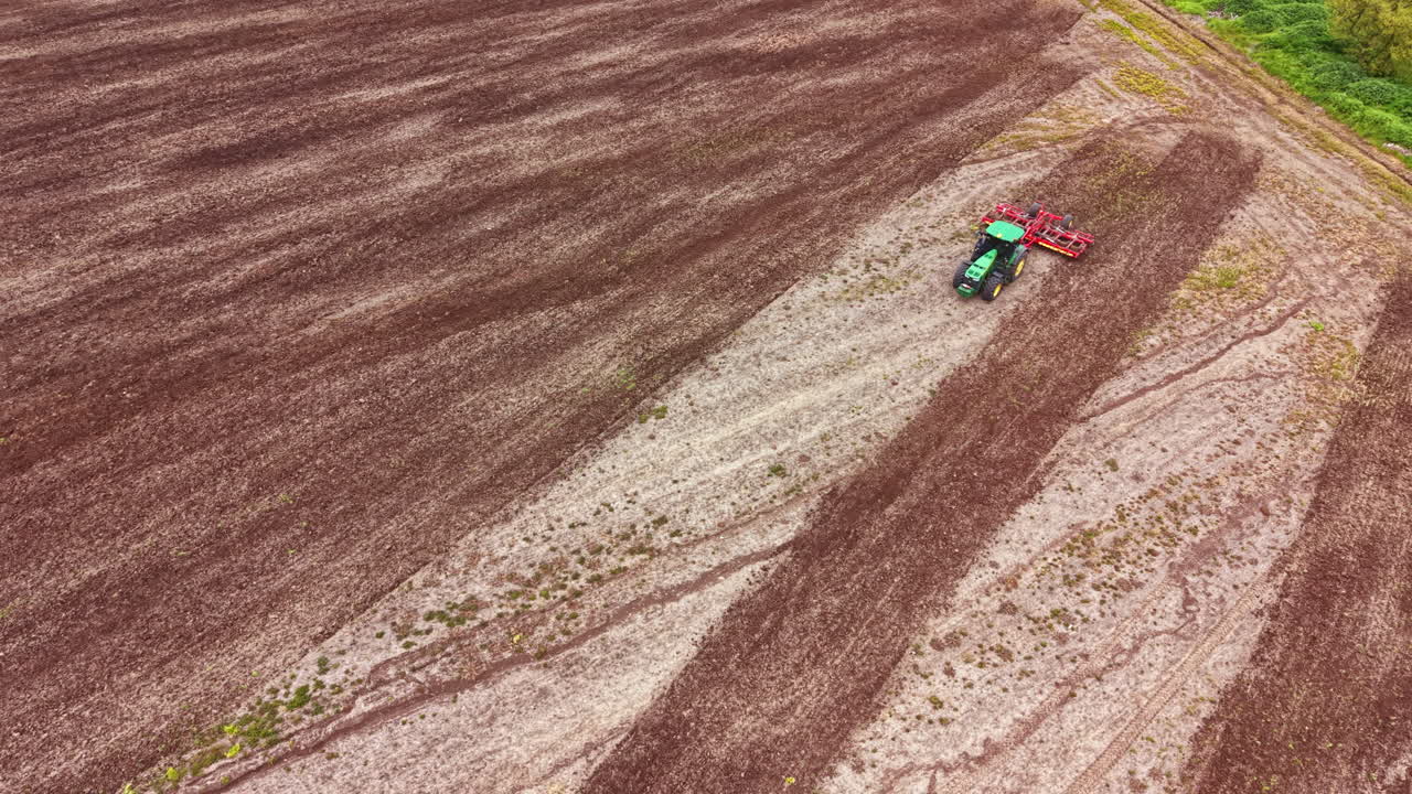 Trattore al lavoro in un campo durante la stagione della semina in una zona rurale con campi coltivati e alberi sullo sfondo.