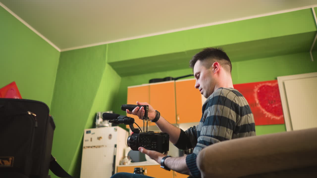 Man reaches for microphone from equipment bag in home office, green walls and cozy setting with camera gear, lens, and smartphone visible. Focused professional preparing gear for shoot