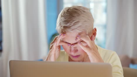 mujer anciana sosteniendo la cabeza con las manos mientras está sentada y trabajando con una computadora portátil