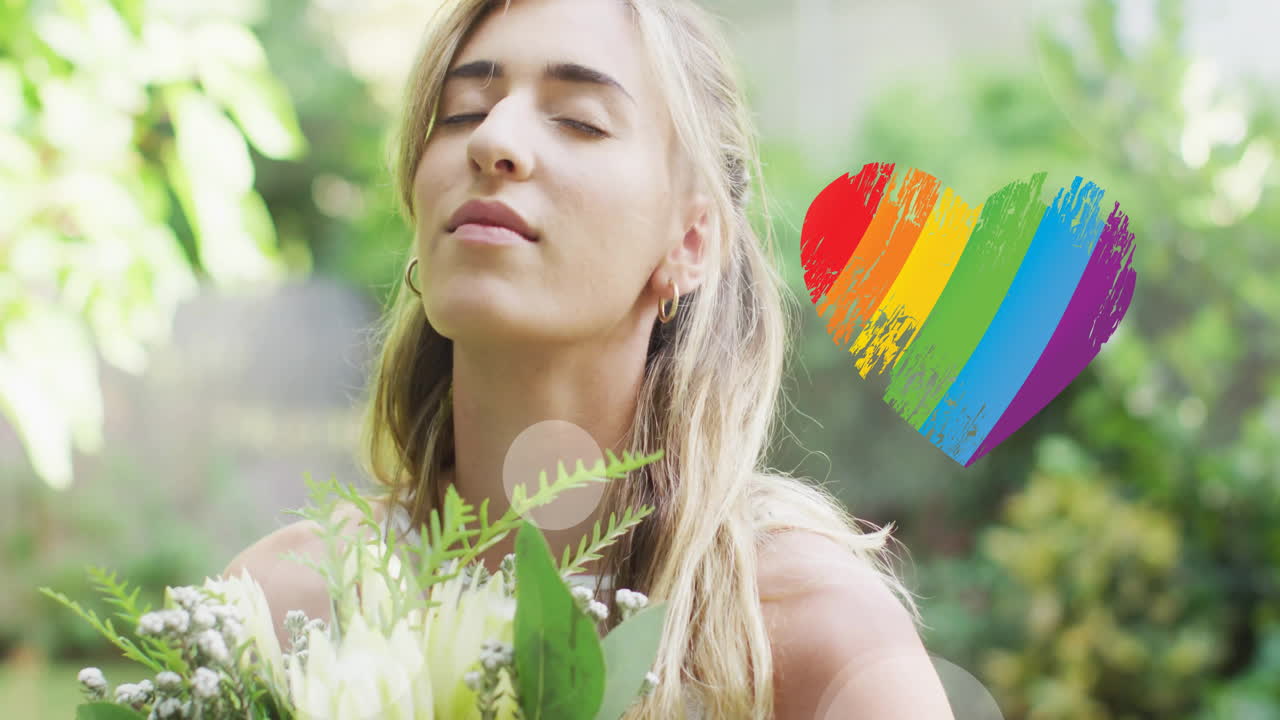 Animation of rainbow heart over caucasian woman wearing wedding dress and holding flowers