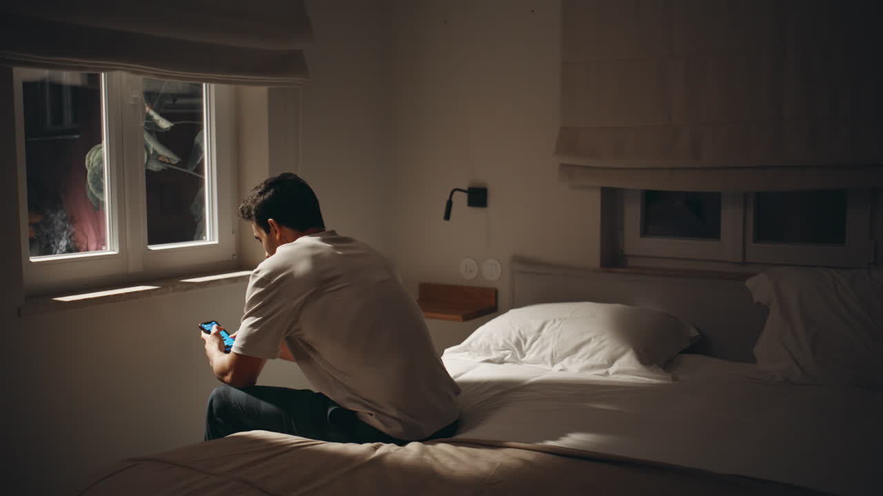 Guy browsing cellphone late evening in dark bedroom. Sleepless man on night bed