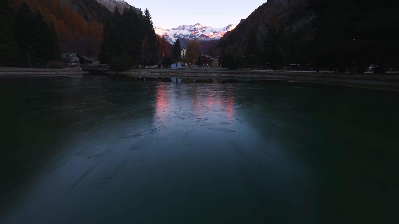 Veduta aerea al tramonto sul lago di Gressoney con Alpi innevate, luci del villaggio e acque serene e riflettenti