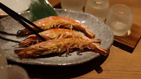 close-up of chopsticks picking up grilled shrimp from a stone plate with decorative leaf and glasses.