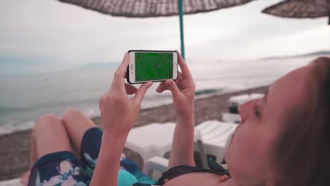 woman in vacation on sunbed, near ocean and vertical mobile phone with green screen, alpha channel and tracking marks. video mock up of smartphone and summer concept.