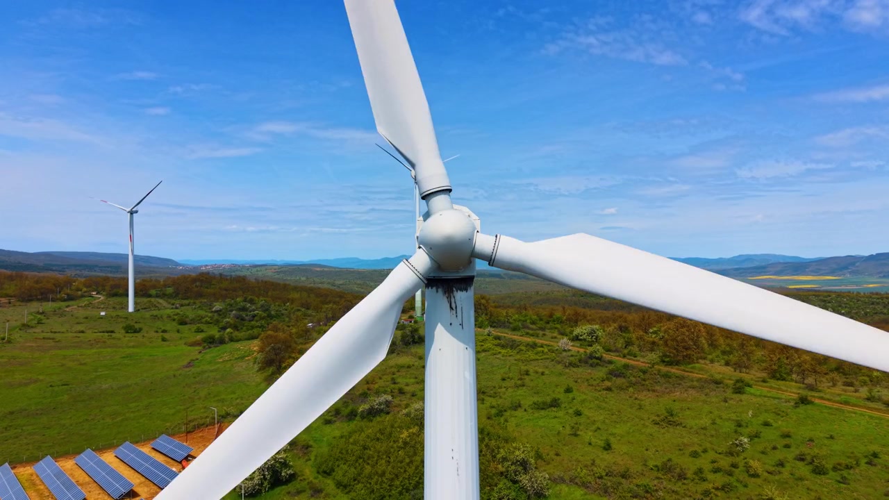 Vista frontale del mozzo del rotore e delle pale di una turbina eolica. Collettori fotovoltaici giacciono su pendii erbosi sotto generatori aggiuntivi e colline distanti.
