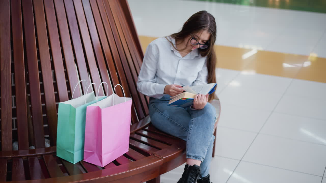 estudiante absorto en un libro, sentado en un banco de madera, bolsas de compras al lado, bajo el brillo brillante de las luces del centro comercial que se reflejan en los azulejos brillantes