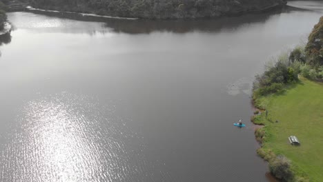 Aerial-birds-eye-view-shot-from-up-high-a-man-on-a-blue-kayak-paddles-out-into-the-middle-of-a-lake