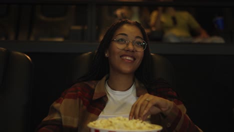 African-American-woman-sitting-in-armchair-watching-a-comedy-at-the-cinema,-taking-popcorn