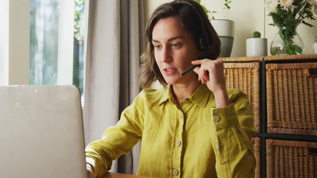 sección media de una mujer caucásica hablando por teléfono con auriculares usando una computadora portátil trabajando desde casa