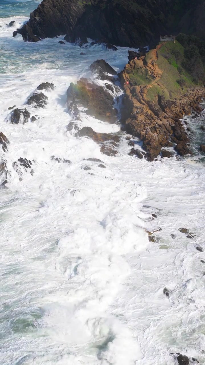 Potenti onde oceaniche si infrangono contro scogliere rocciose sotto la luce solare intensa, creando una scena costiera dinamica e drammatica.