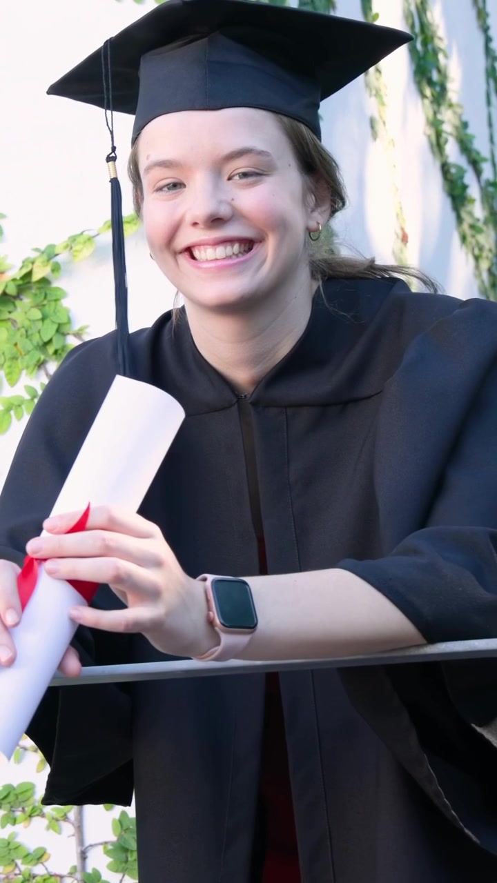 Vertical video: Recording graduate at ivy wall in cap and black gown, turning and showing diploma