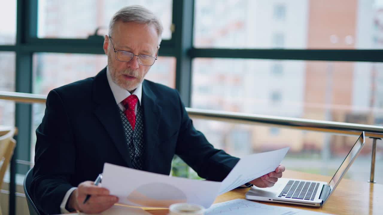 Serious businessman at the table. Mature man in black suit working with documents near the window in the office.