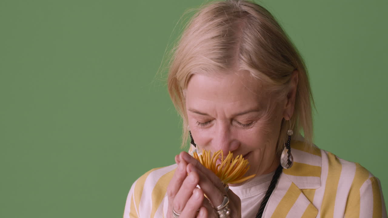 줄무늬 셔츠와 액세서리를 입은 파란 눈을 가진 금발의 성숙한 여인은 해바라기 냄새를 맡고 녹색 배경에서 카메라를 보고 웃고 있습니다.