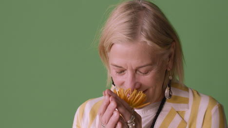 blonde mature woman with blue eyes dressed in striped shirt and accessories posing smelling a sunflower and smiling at camera on green background