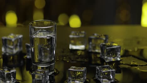 barman pour frozen vodka from bottle into shot glass. ice cubes against shiny gold party background