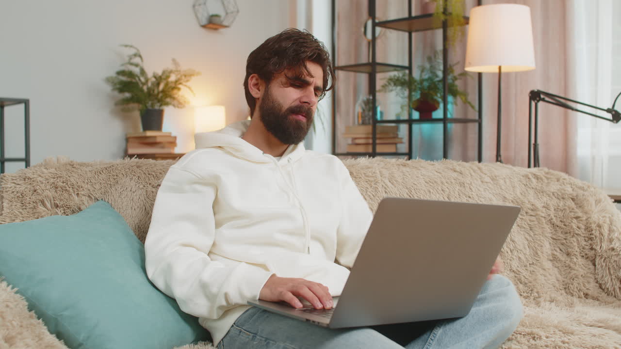 Upset young indian man freelancer sitting on sofa at home using laptop having anxiety and stress