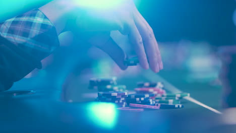 Defocused-Hands-Of-A-Casino-Player-Holding-Chips-On-A-Wooden-Table-With-Green-Bright-Lights-Background