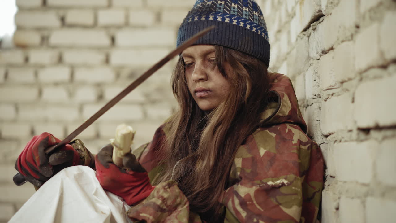 Young girl dressed in camo jacket and knit cap sits against rough brick wall shaping stick with large cutlass, concentrating with red gloves and determination
