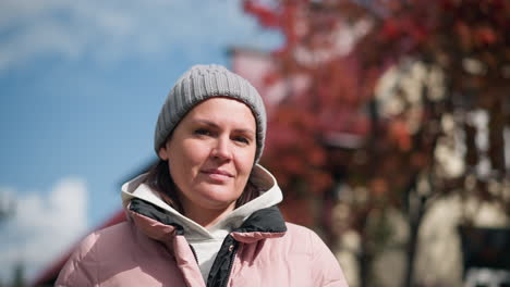 young woman in pink jacket and grey beanie, looking thoughtful, tilting head with a gentle blink, outdoors with blurred autumn trees and house in background under clear sky