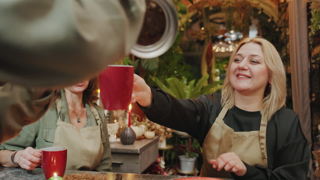 To blomsterhandlere nyder kage og kaffe ved et rustikt bord, butiksejere skåler med røde krus blandt stearinlys og grønt, varm sæsonbestemt indretning og venlig samtale i en intim blomsterbutik