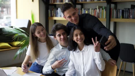 Estudiantes-Europeos,-Un-Grupo-De-Cuatro-Se-Toman-Selfies-En-La-Biblioteca-Del-Colegio-O-Universidad.-Grupo-De-Cuatro-Parados-Frente-A-Estanterías