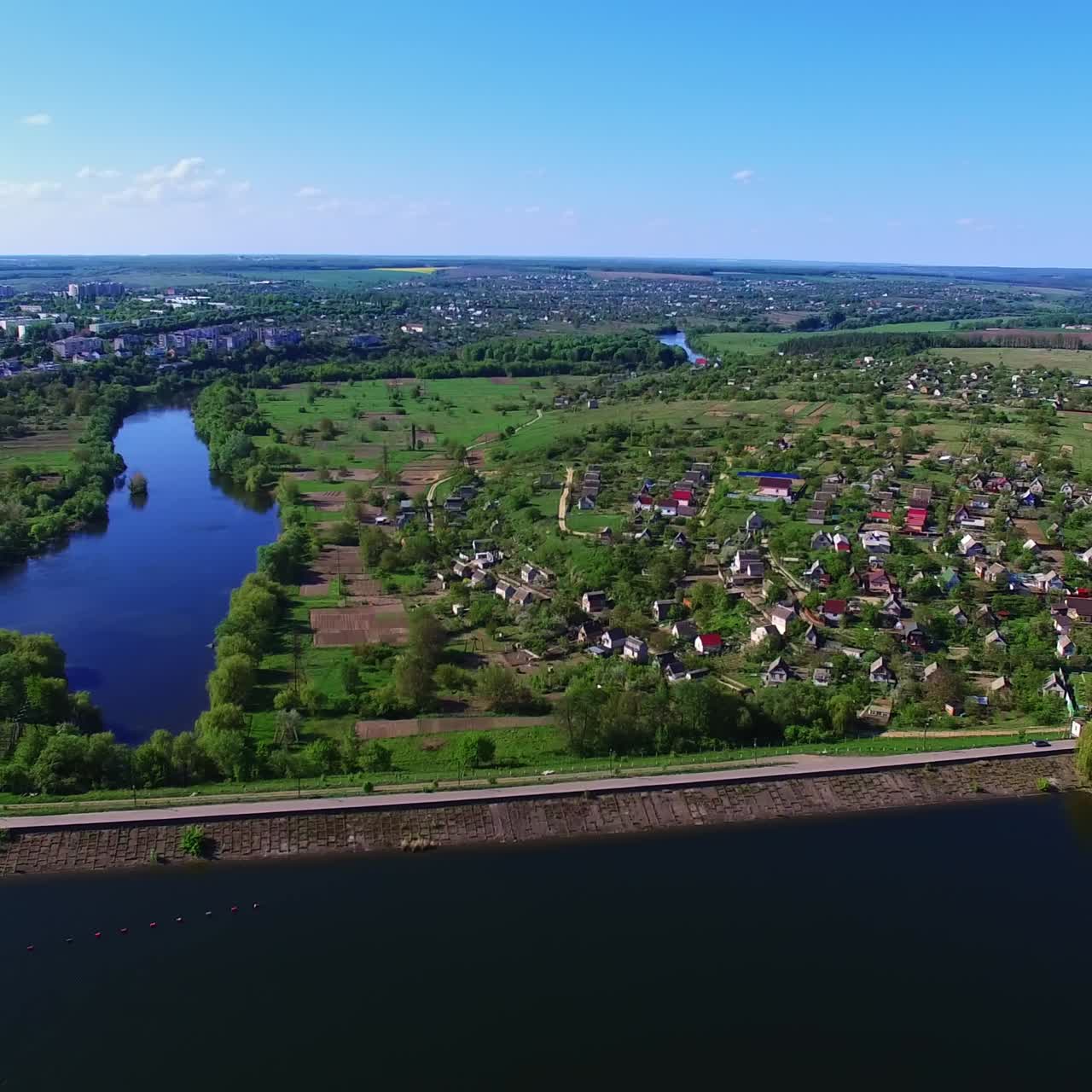Approaching the beautiful village dwelling on the bank of blue river. Green landscape under clear sky at backdrop. Top view