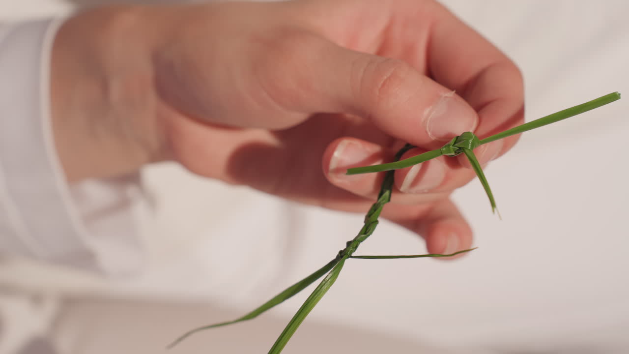 Tangan Kaukasia menganyam simpul rumput sebagai kenang-kenangan pernikahan, close-up intim jari pengantin wanita mengikat simpul halus di bawah cahaya matahari lembut, simbol keluarga dan pernikahan buatan tangan, latar belakang minimalis pedesaan, suasana tenang.
