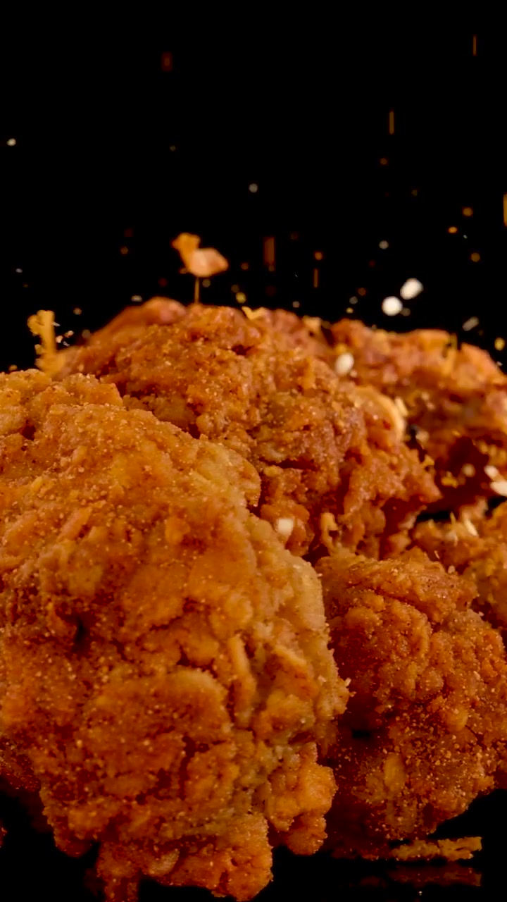 Pezzi di pollo impanati e dorati vengono conditi con spezie che cadono su uno sfondo scuro.