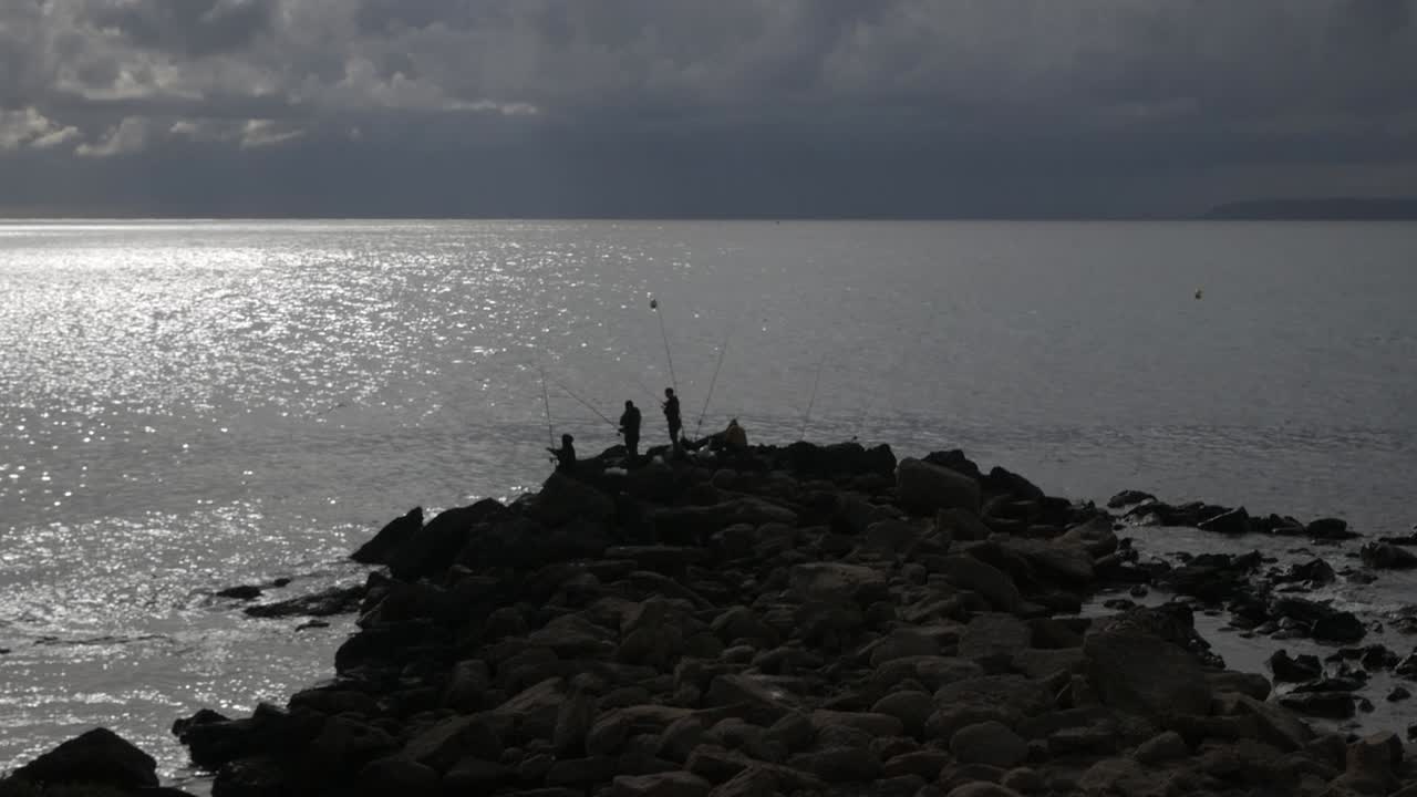 Fishermen on rocky shore in Cabo De Huertas, Alicante, under cloudy skies at sunset
