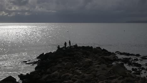 fishermen on rocky shore in cabo de huertas, alicante, under cloudy skies at sunset