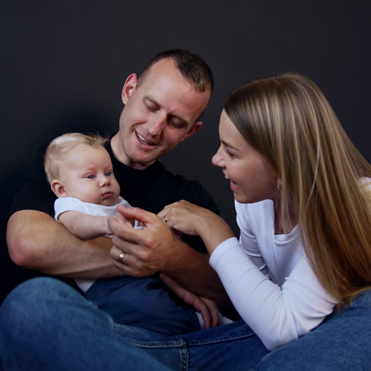 Father holds his tiny serious baby boy. Dad waves kid's hands and mother talks and smiles to her son