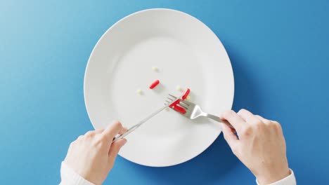 Video-of-hands-holding-knife-and-fork-over-dinner-plate-of-various-pills-on-blue-background