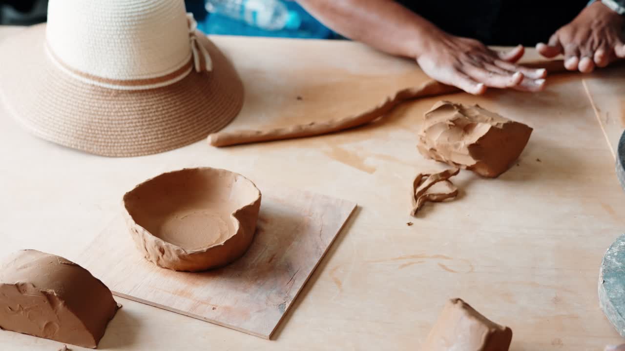 Hands roll a clay coil and shape a rustic bowl using hand-building pottery techniques on a sunlit table, with a straw hat nearby.