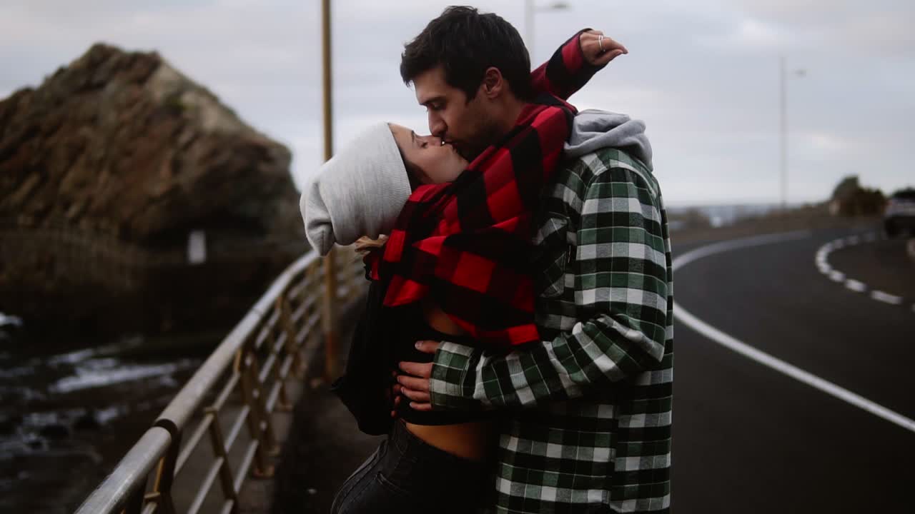 hombre pensativo mira fijamente al océano - pareja joven mostrada desde el lado de pie en la carretera costera. chica linda y elegante viene a su novio para besarlo. clima nublado