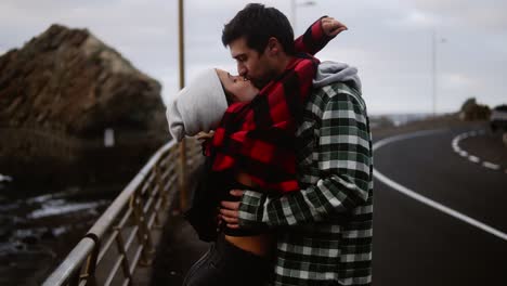 Thoughtful-man-gazes-at-ocean---Young-couple-shown-from-the-side-standing-on-coastline-road.-Cute,-stylish-girl-comes-to-her-boyfriend-to-kiss-him.-Cloudy-weather