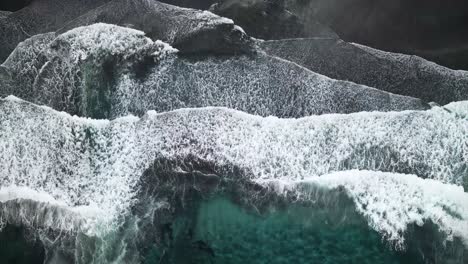 aerial overhead view capturing powerful ocean waves crashing on the black sand beach of tjørnuvík, highlighting the dramatic contrast between dark sand and turquoise water in the faroe islands