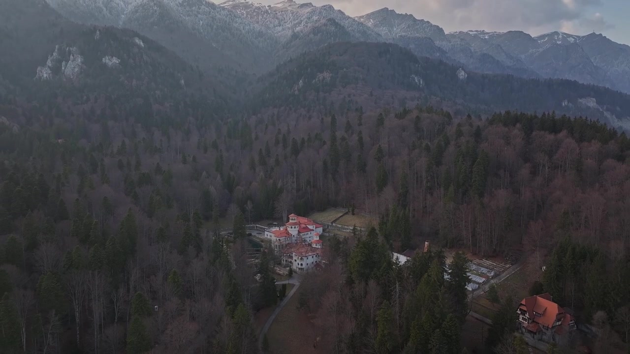 Inquadratura aerea dall'alto con movimento tilt-up che rivela il Rifugio di caccia Foisor immerso in una fitta foresta di pini con cime montuose innevate a Sinaia, Romania