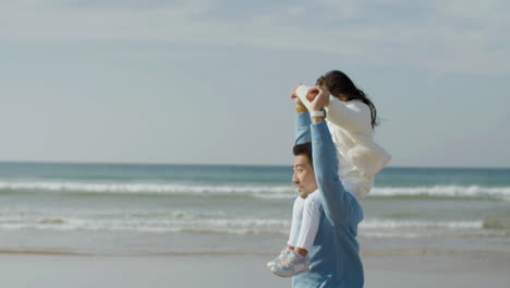 Feliz-Padre-Japonés-Caminando-Por-La-Orilla-Del-Mar-Con-Su-Hija-Sobre-Sus-Hombros