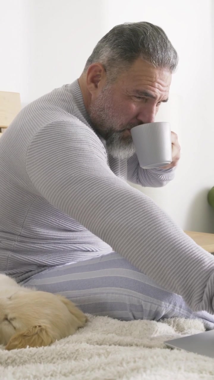 Video vertical: Hombre en pijama inclinado hacia adelante, extendiéndose para tomar un sorbo de una taza después de ver el portátil en el dormitorio.