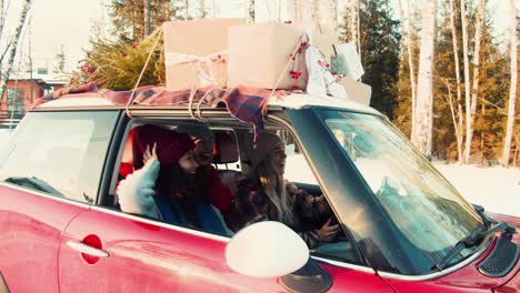 three young happy multiethnic women friends drive red car with christmas presents on top along snowy road slow motion.