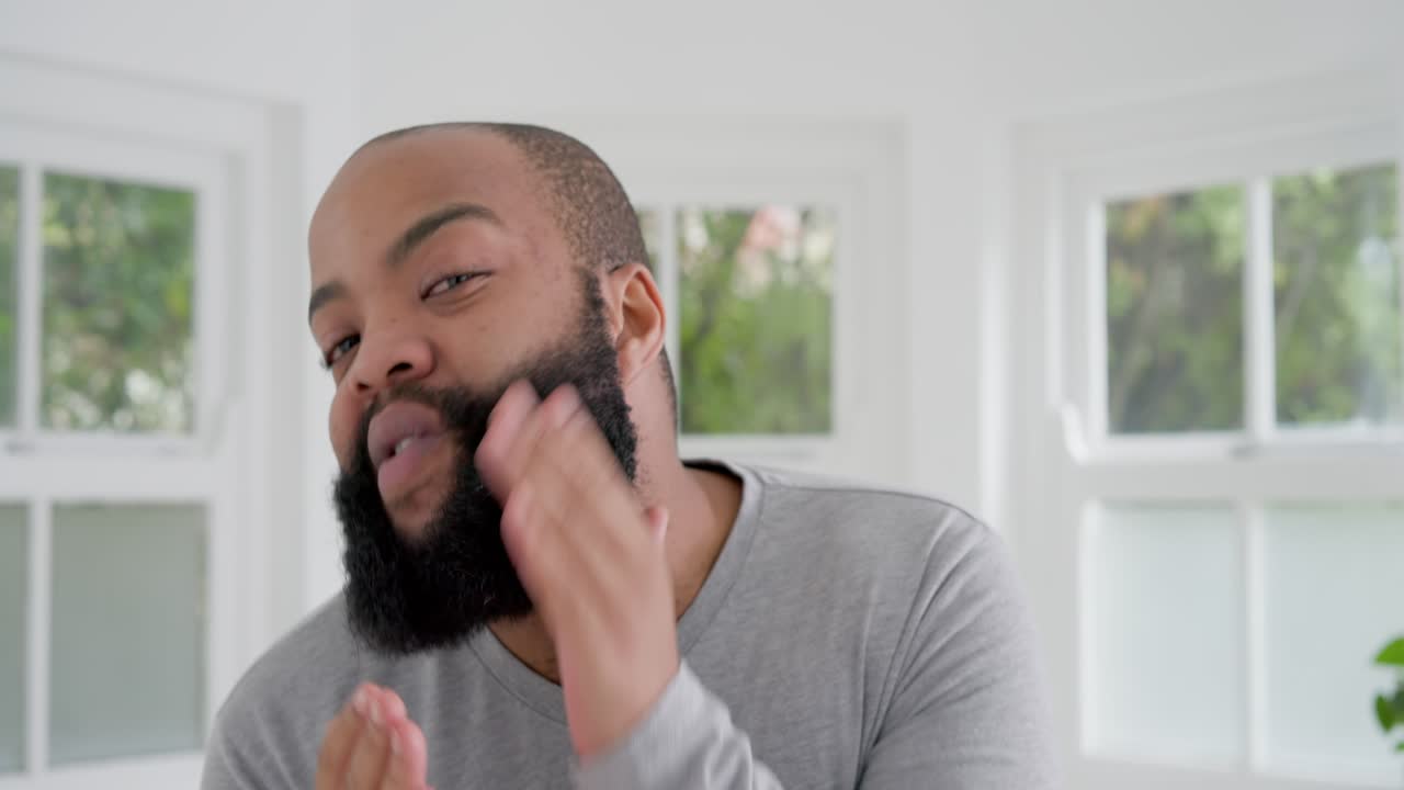 African American man grooming beard in white room by windows, bringing hand to cheek, checking face