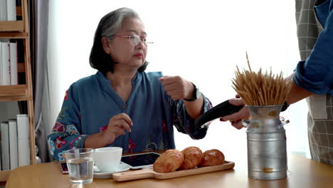 Active-senior-Asian-woman-working-in-a-coffeeshop-using-a-mobile-device,-smartwatch-for-NFC,-QR-code-scanning,-contactless-payment-to-pay-for-coffee-and-snack