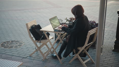 freelancer working on laptop outdoors in cafe, sitting at table with plants, surrounded by wooden chairs, and a handbag placed nearby, with a paved walkway and buildings blurred in the background